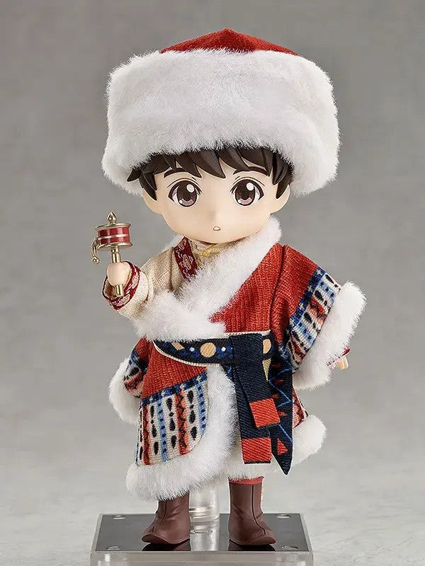 Brown-haired doll in a red fur-trimmed hat and patterned robe, holding a prayer wheel on a display stand.