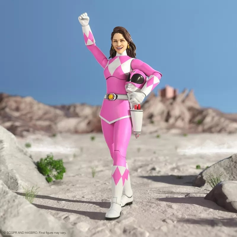 Pink Ranger figure with an unmasked head, raised fist and helmet tucked under one arm in a rocky setting.