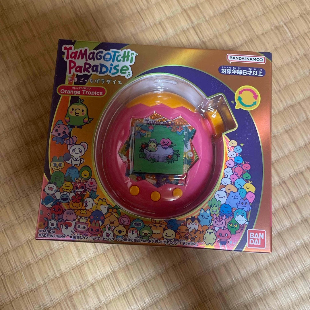 A boxed pink and orange Tamagotchi Paradise device in retail packaging on a wooden floor.