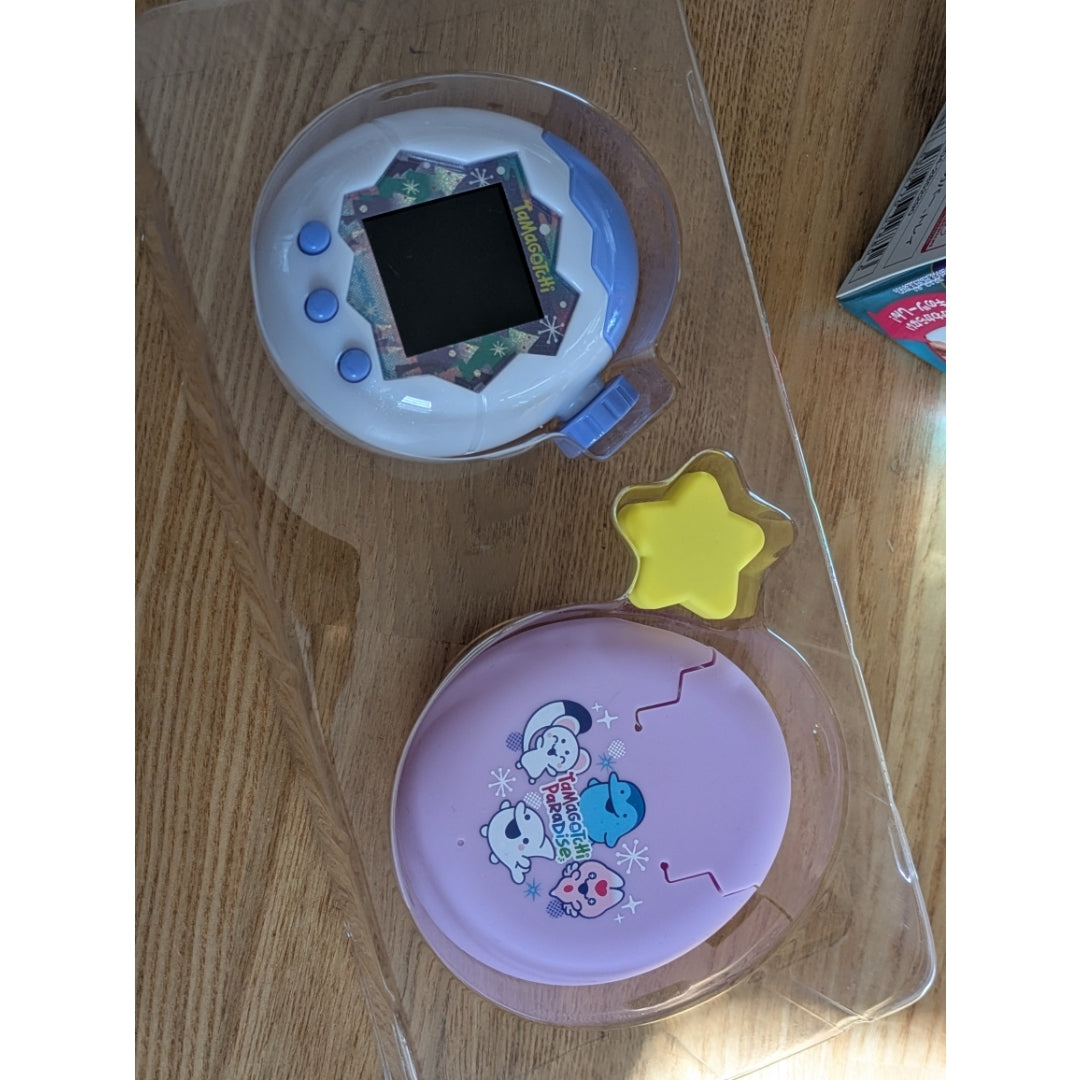 Two Tamagotchi Paradise devices in clear plastic trays on a wooden surface.
