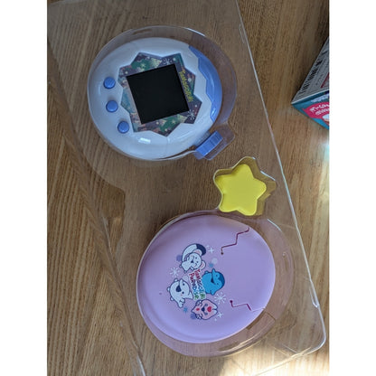 Two Tamagotchi Paradise devices in clear plastic trays on a wooden surface.